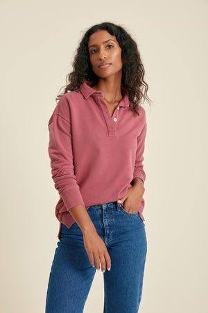 Woman wearing a pink long-sleeve polo polo and blue jeans against a beige background