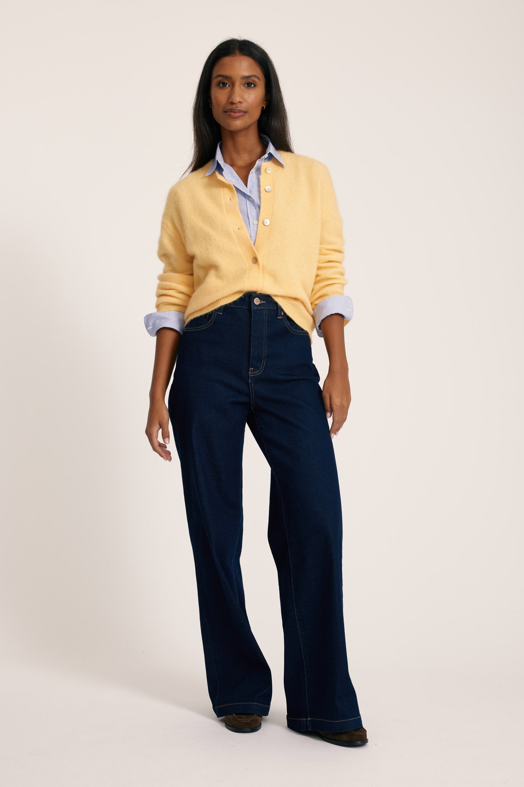 Woman wearing a yellow cardigan, light blue shirt, and dark blue jeans on a white background
