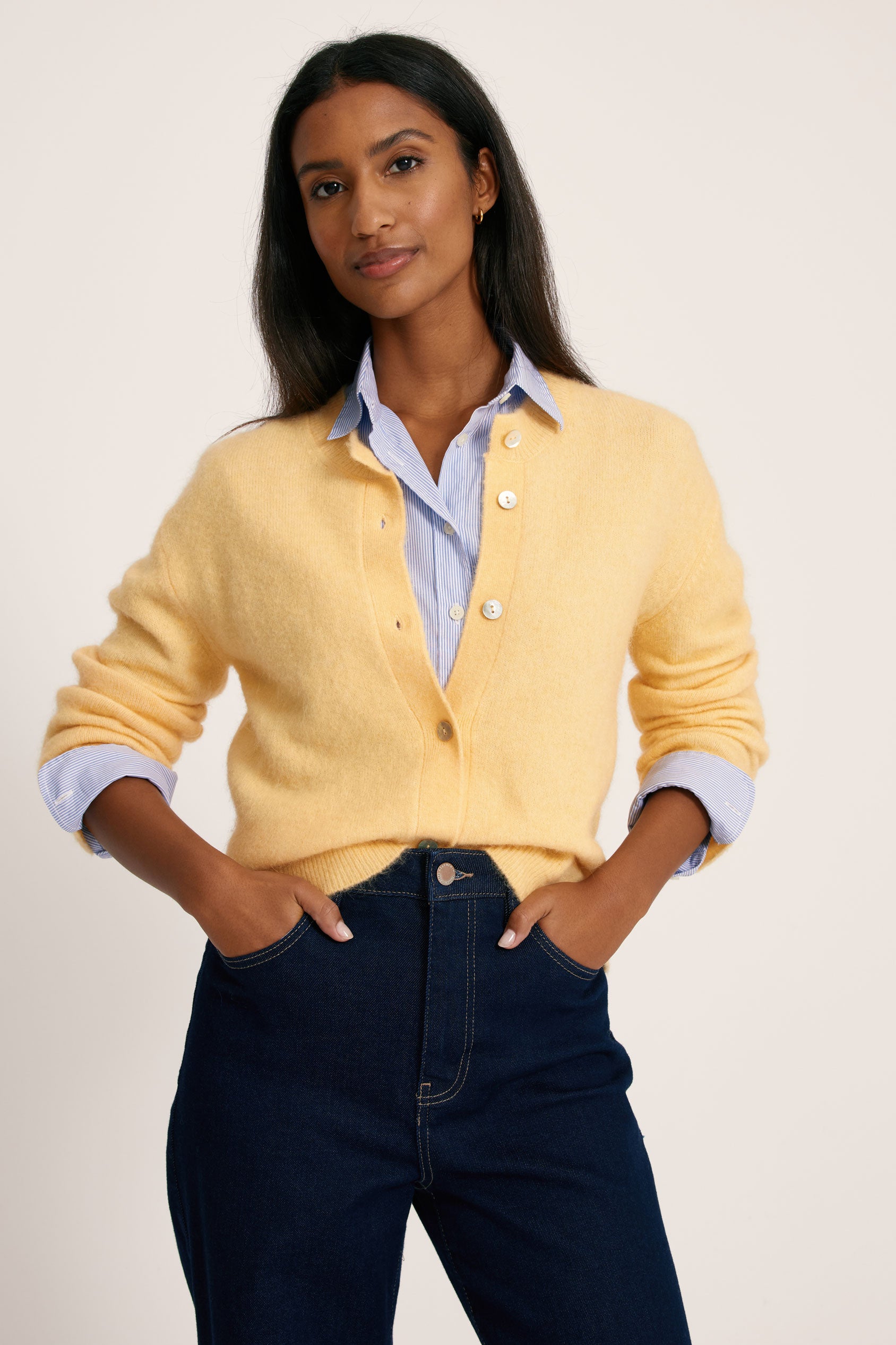 Woman wearing a yellow cardigan over a blue shirt with rolled-up sleeves, standing against a plain background.