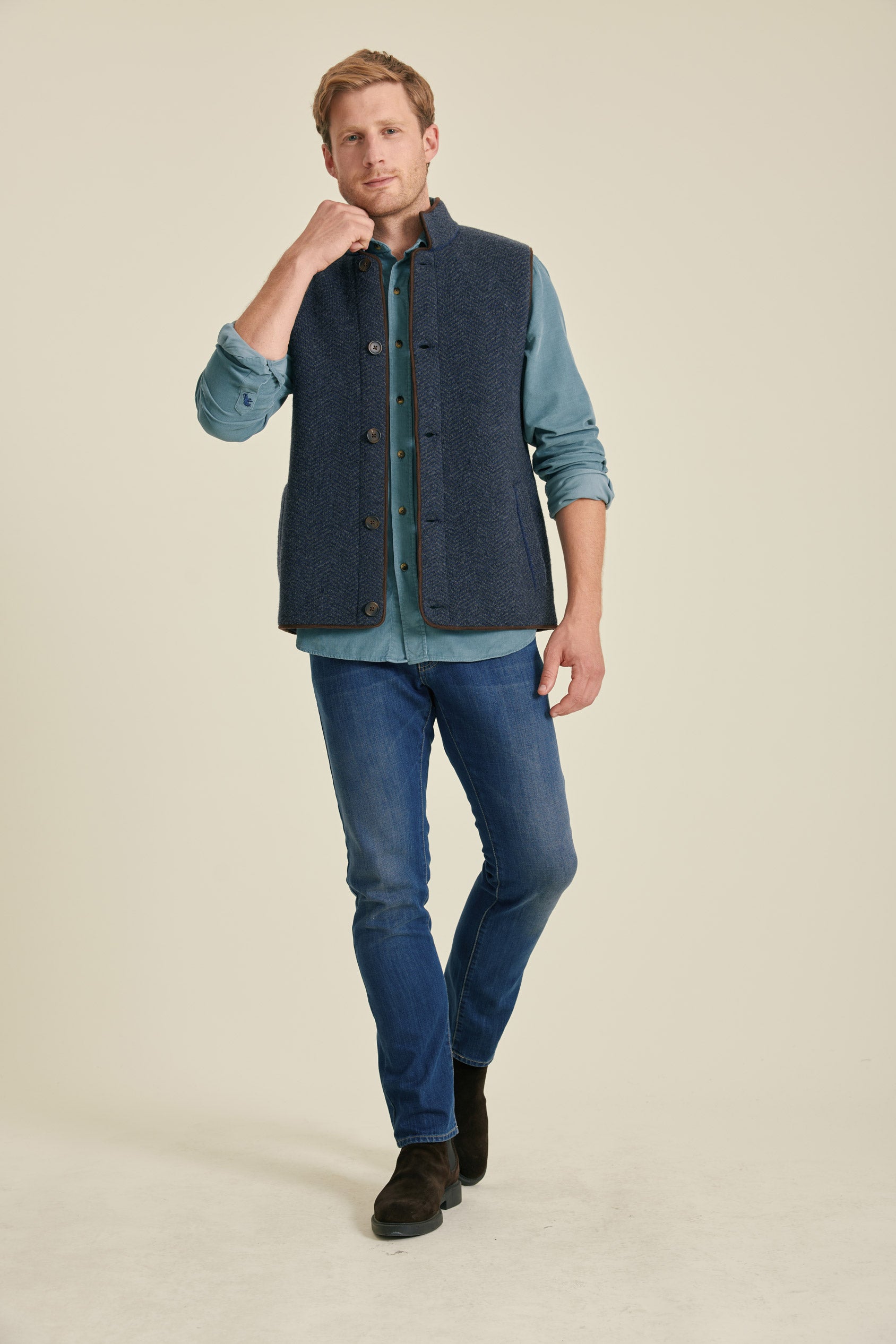 Man wearing a blue shirt, dark vest, and jeans on a beige background