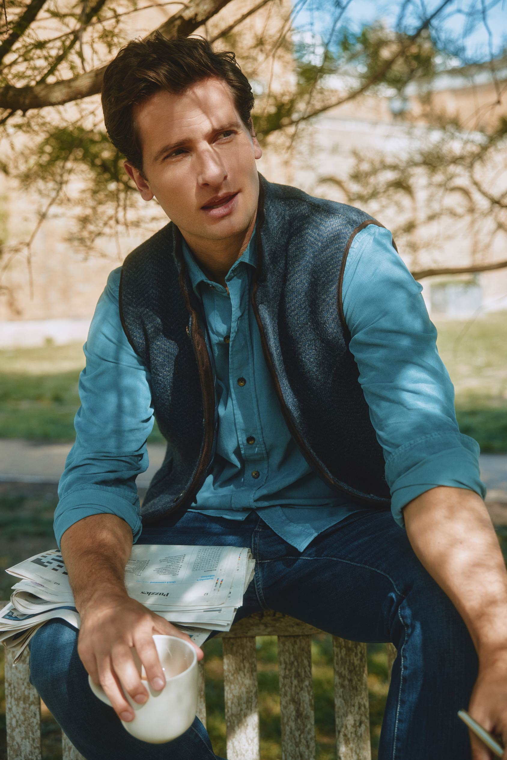 Man sitting outdoors holding a newspaper and a cup, wearing a blue shirt and dark vest.