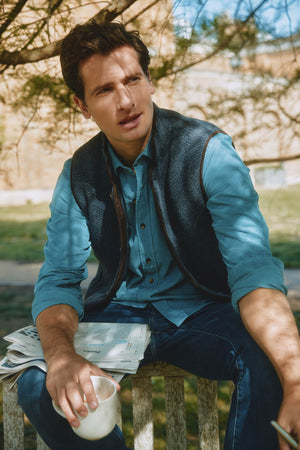 Man sitting outdoors holding a newspaper and a cup, wearing a blue shirt and dark vest.