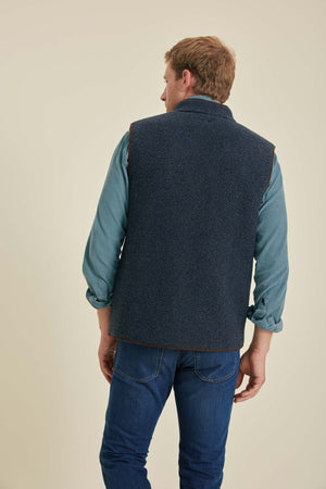 Man wearing a navy blue vest over a light blue shirt and jeans on a beige background