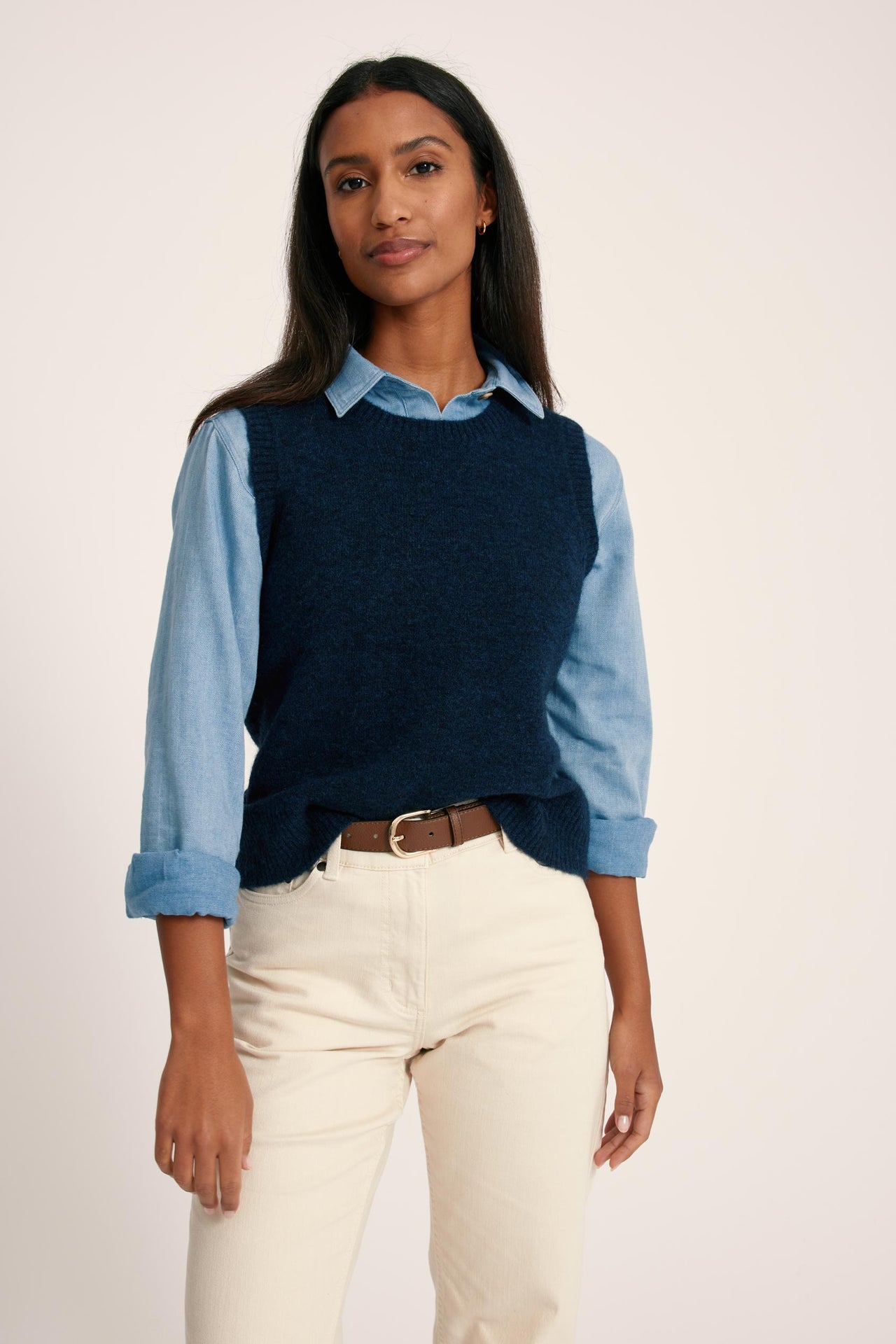 Woman wearing a navy sweater over a light blue shirt with rolled-up sleeves, paired with white pants on a plain background.
