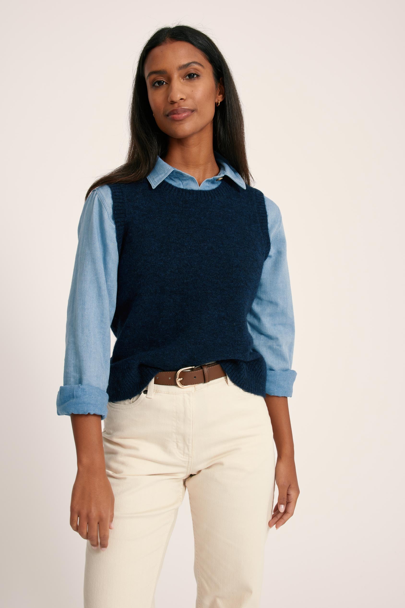 Woman wearing a navy sweater over a light blue shirt with rolled-up sleeves, paired with white pants on a plain background.