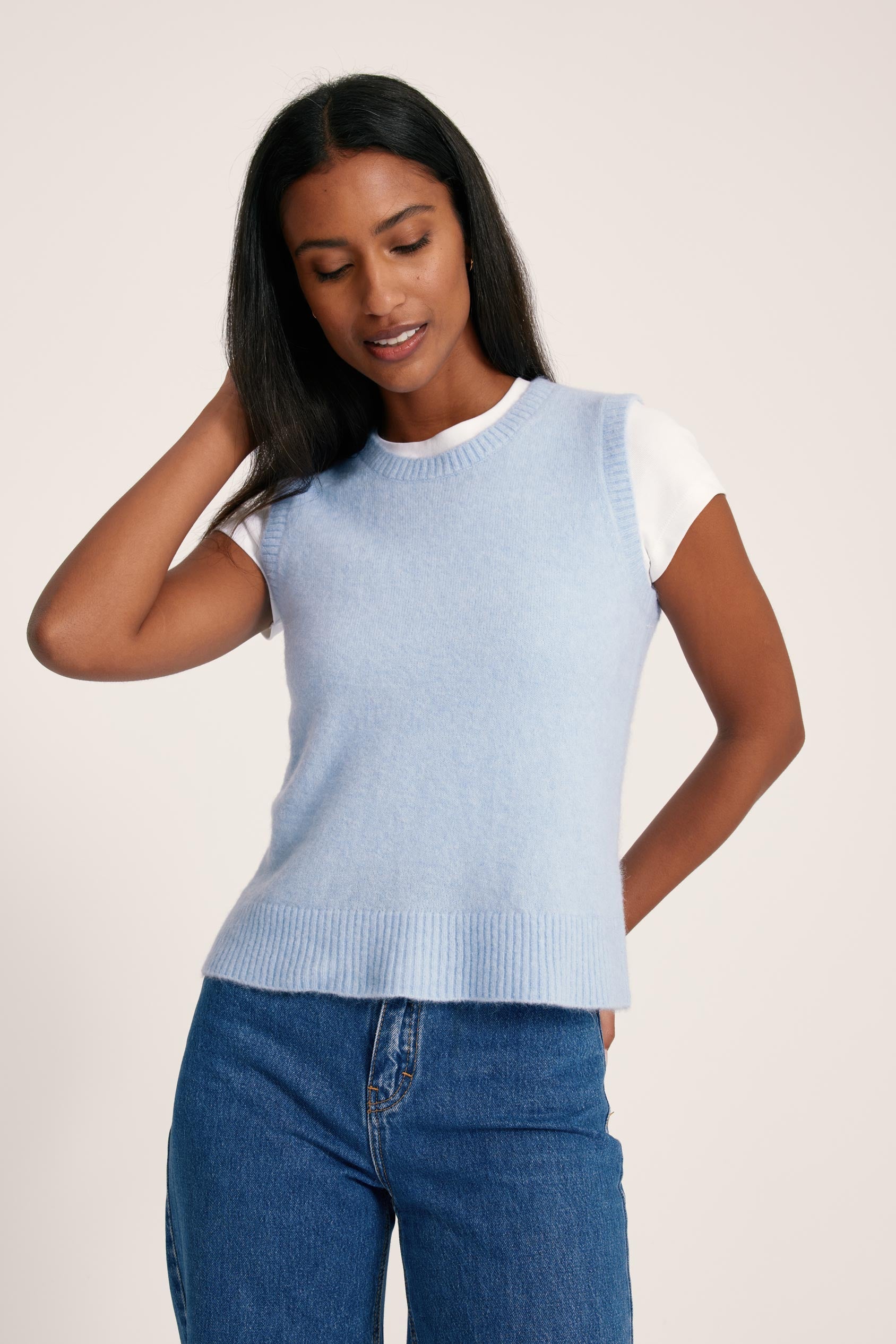 Woman wearing a light blue sleeveless sweater and jeans on a plain background