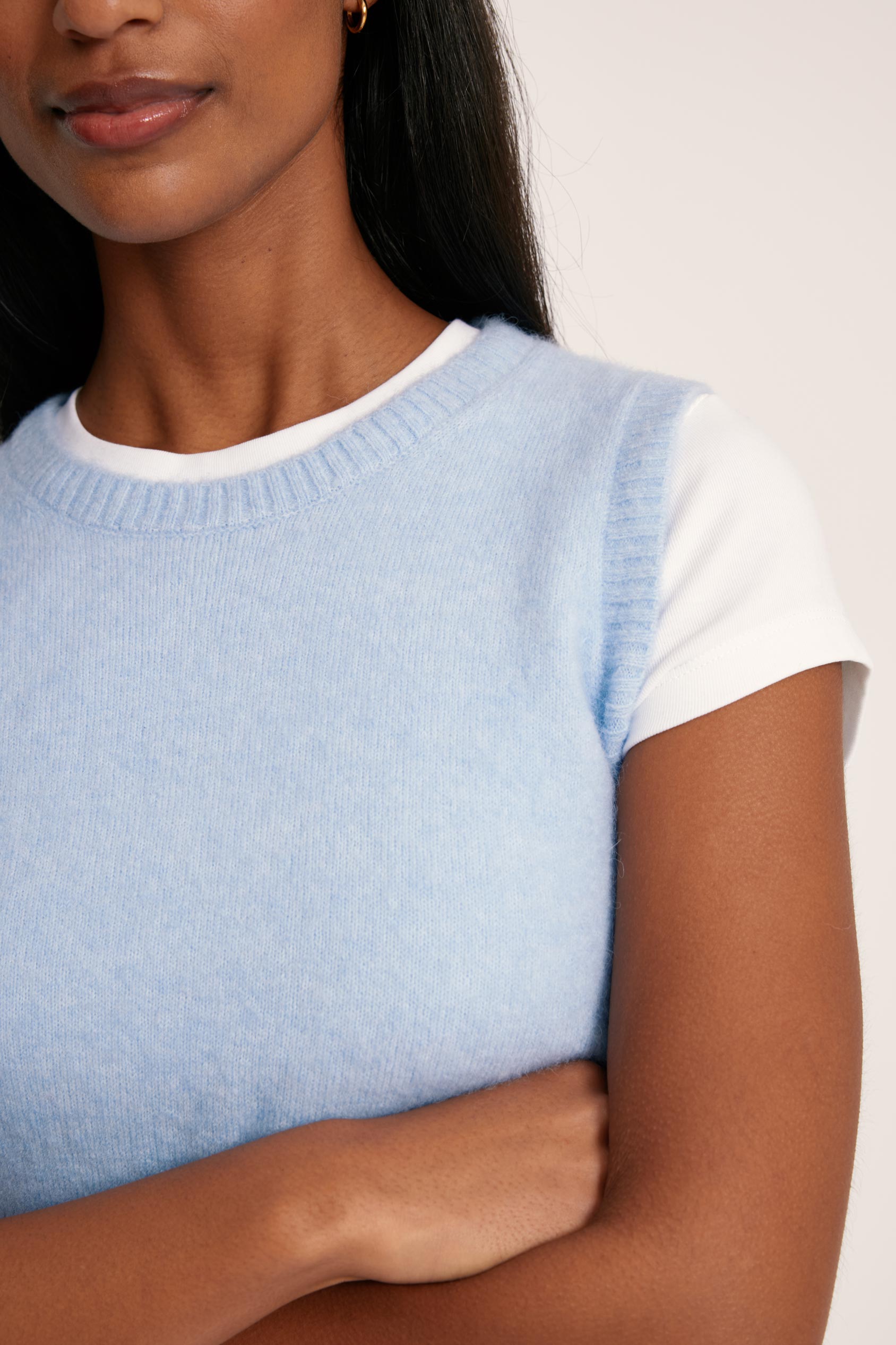 Woman wearing a light blue sleeveless sweater over a white shirt against a plain background