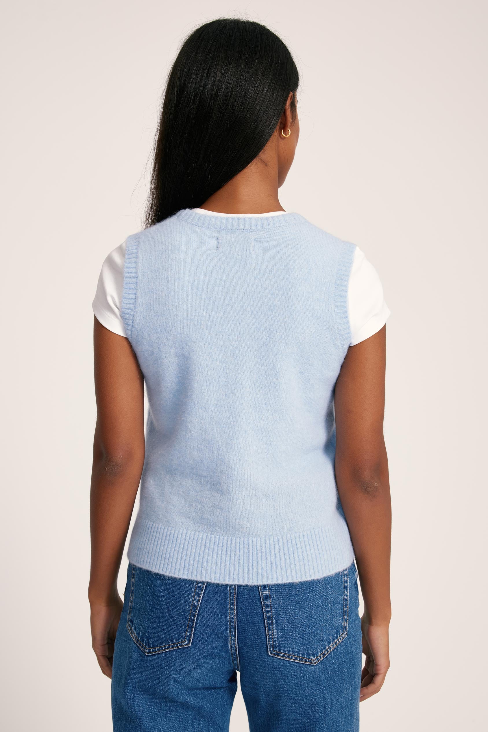 Person wearing a light blue sleeveless sweater over a white shirt with blue jeans on a plain background