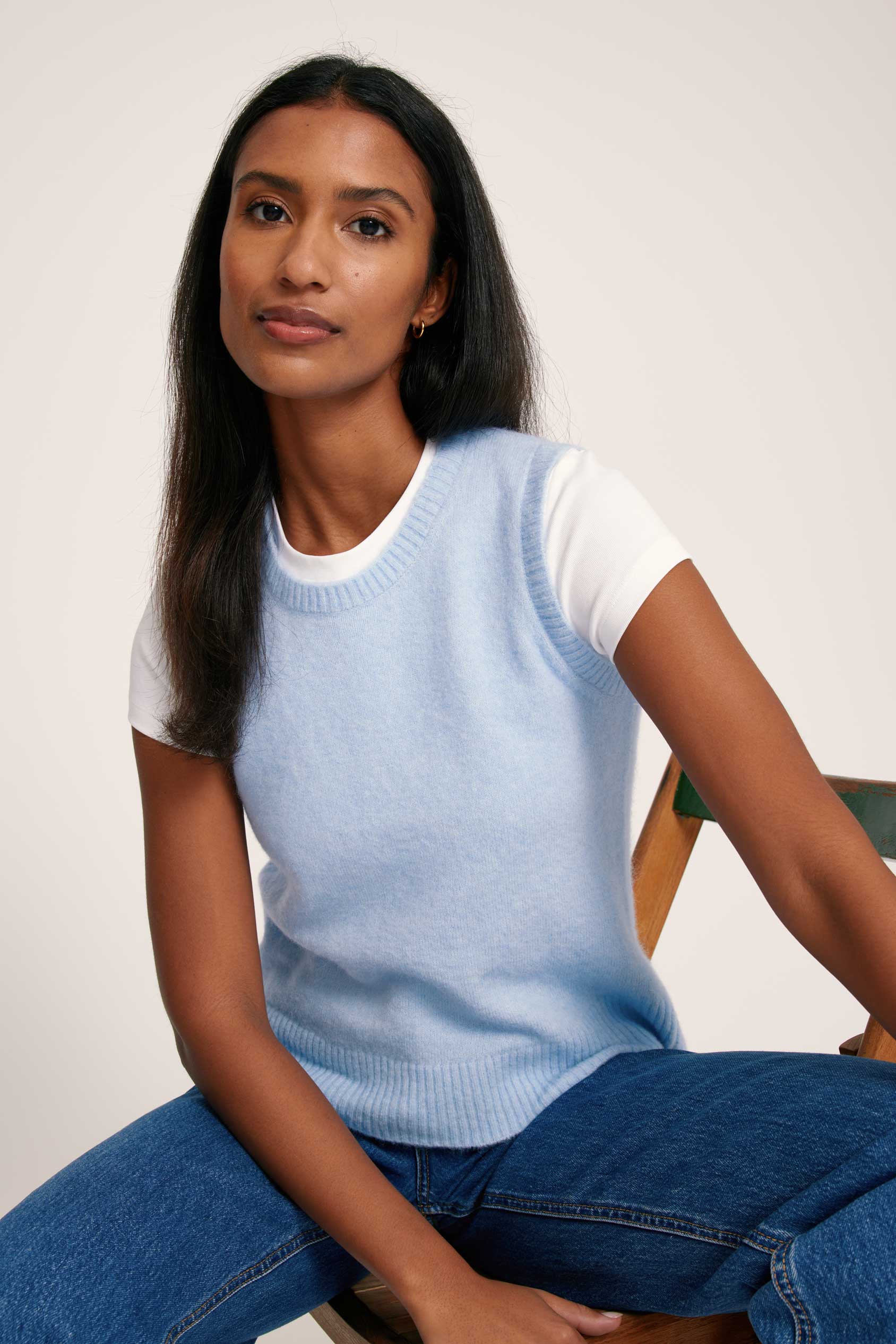 Woman wearing a light blue sleeveless sweater over a white shirt with blue jeans, sitting on a chair.