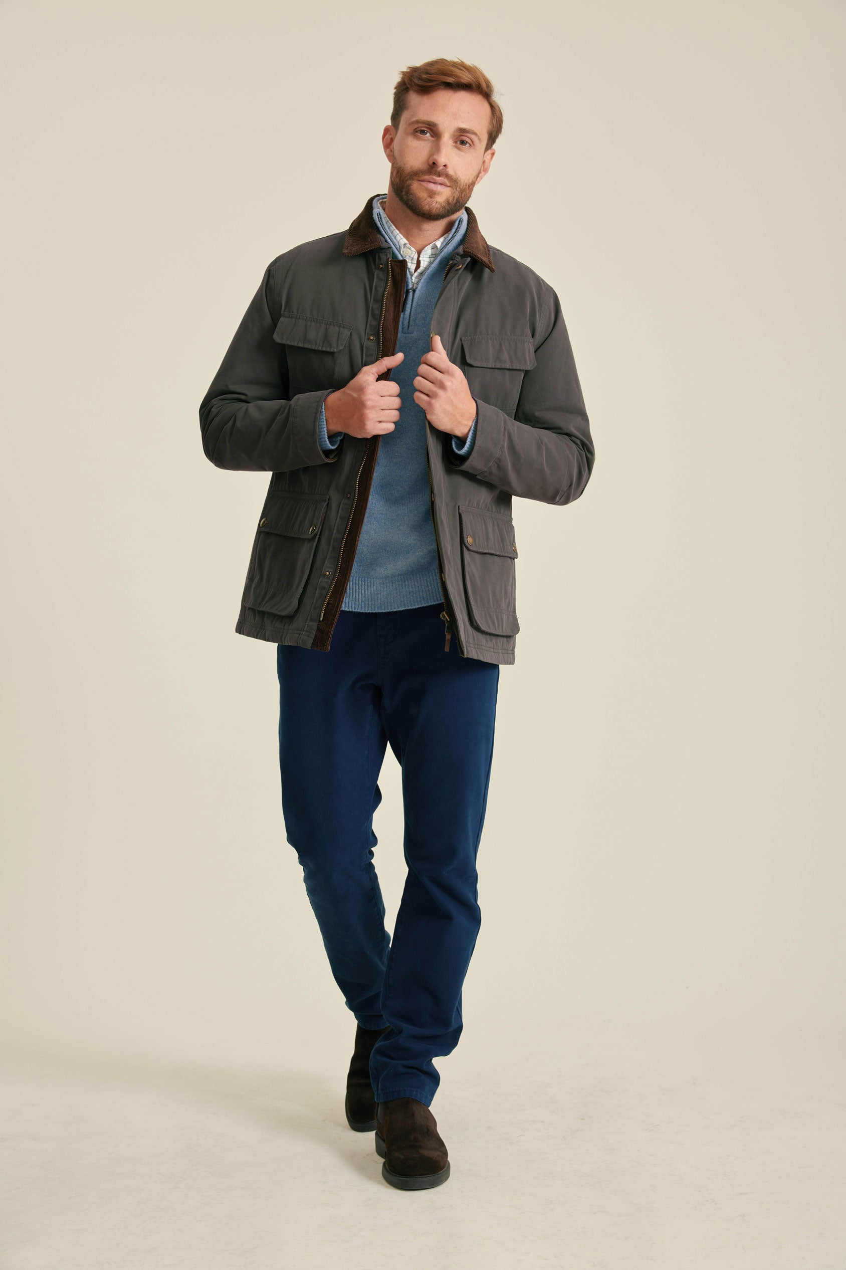 Man wearing a gray jacket, blue shirt, and dark pants on a beige background