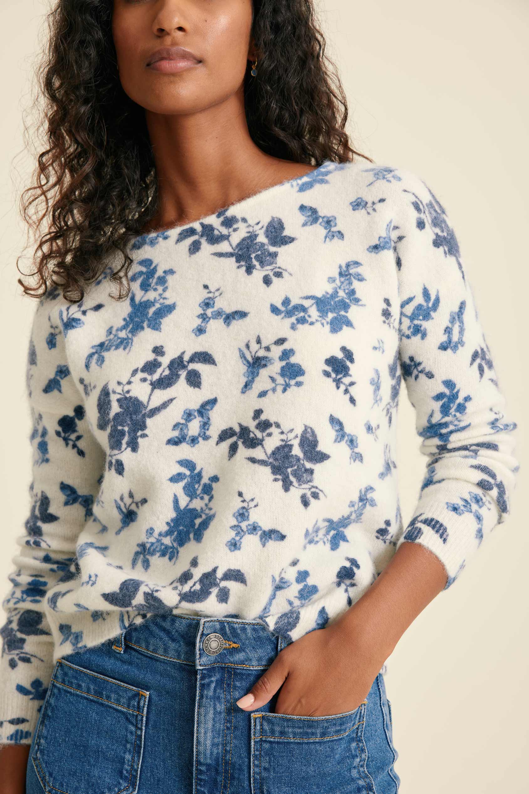 Woman wearing a white sweater with blue floral pattern and blue jeans.