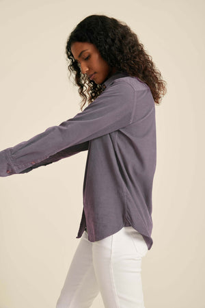 Woman wearing a long-sleeve purple shirt and white pants against a beige background