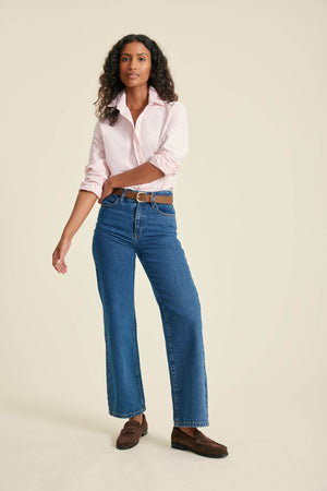 Woman wearing a light pink shirt, blue jeans, and brown shoes on a beige background