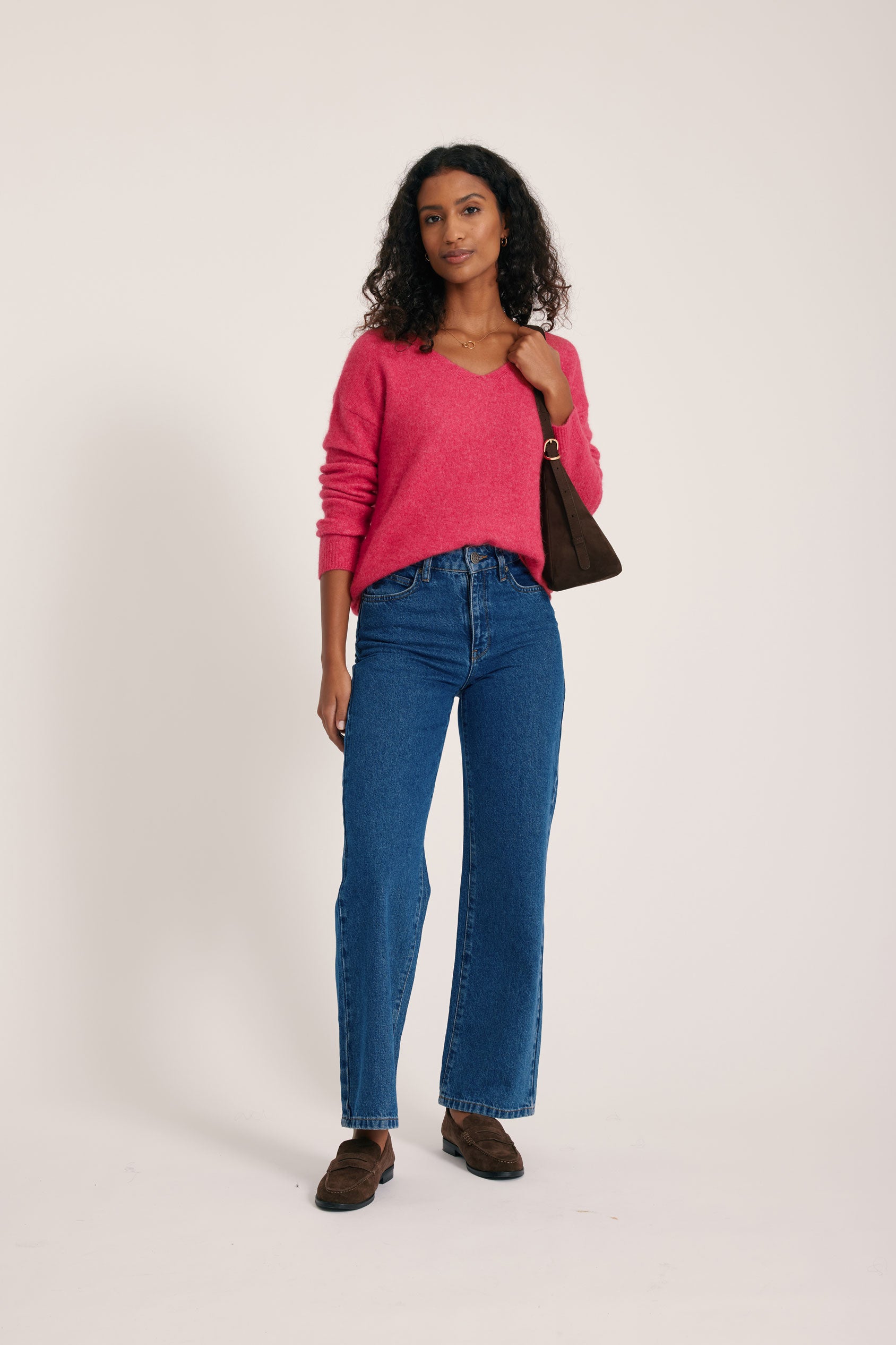 Woman wearing a pink sweater and blue jeans holding a brown handbag on a white background