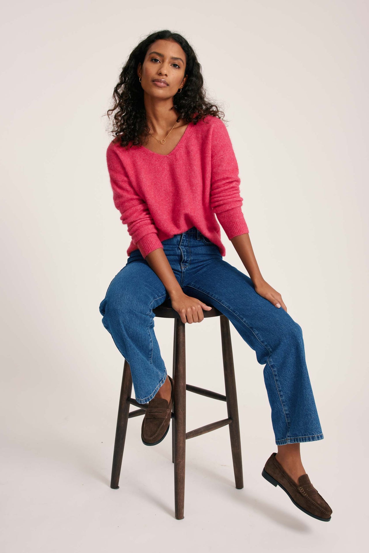 Woman sitting on a stool wearing a pink sweater and blue jeans against a white background