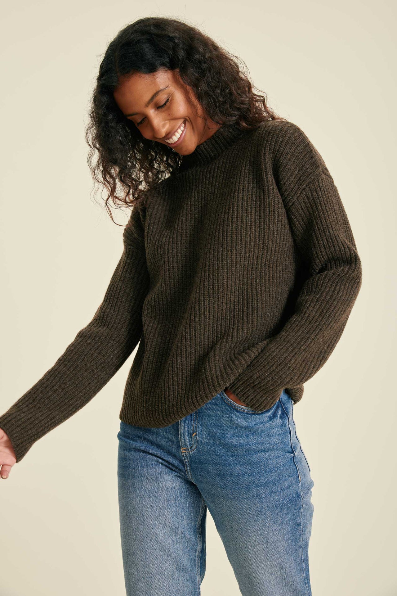 Woman wearing a brown ribbed sweater and blue jeans against a beige background