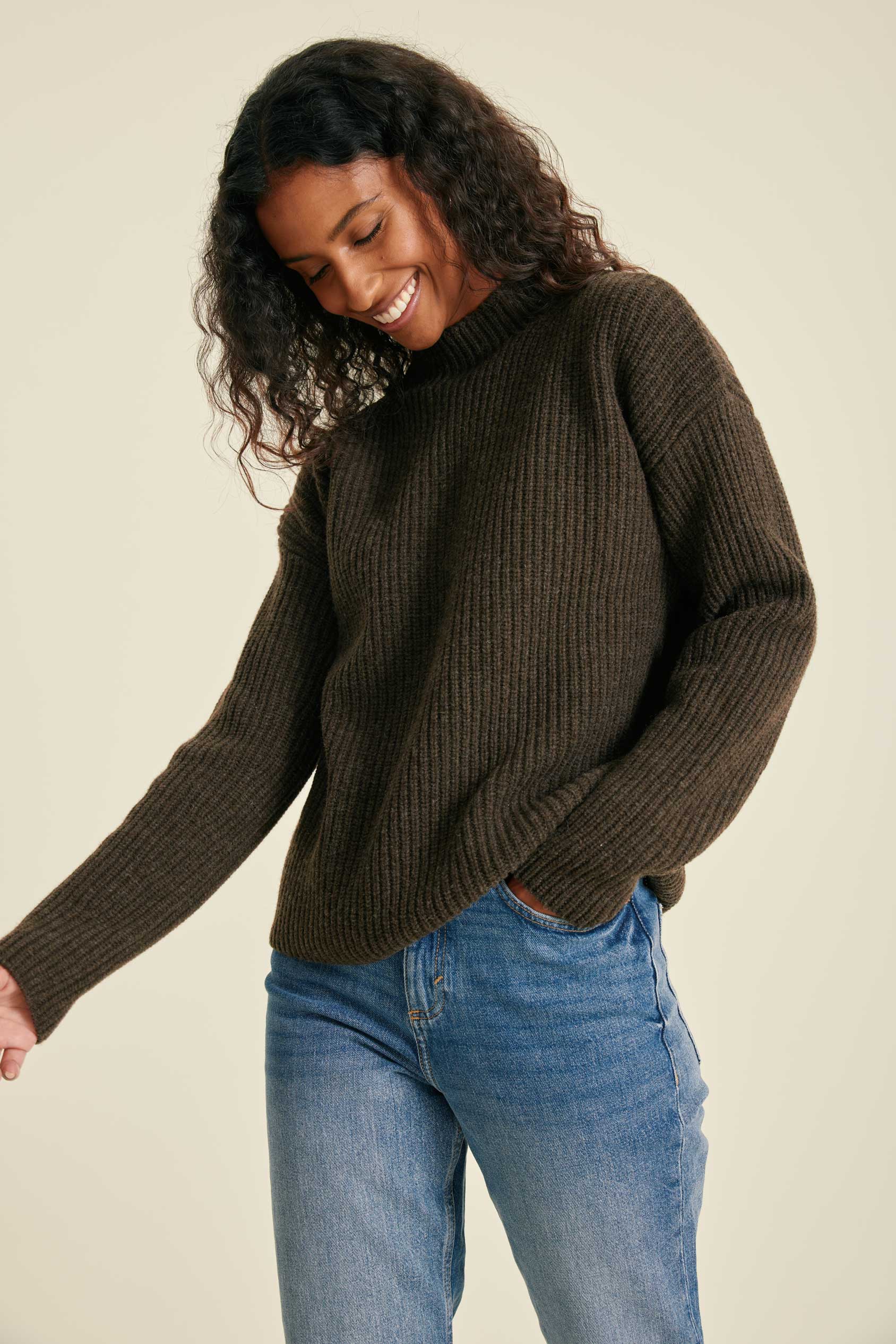 Woman wearing a brown ribbed sweater and blue jeans against a beige background