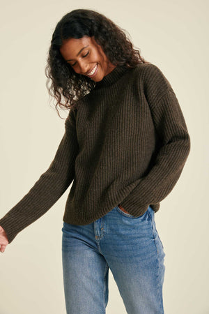 Woman wearing a brown ribbed sweater and blue jeans against a beige background