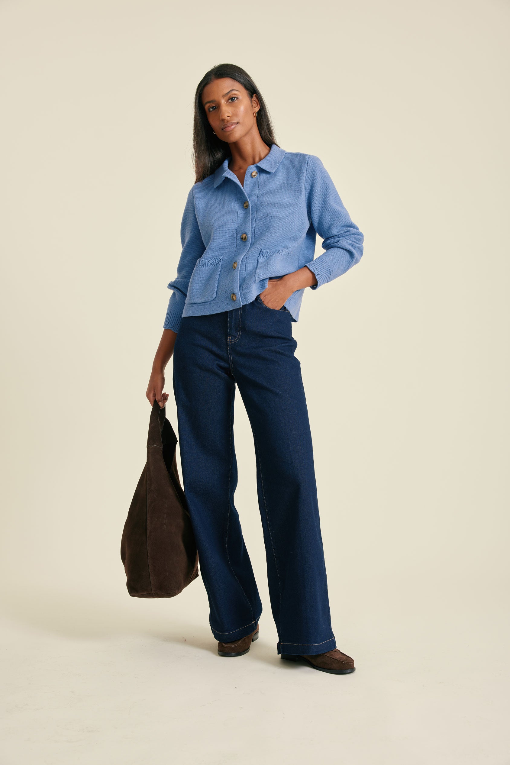 Woman wearing a blue shirt and dark blue jeans holding a brown bag on a beige background