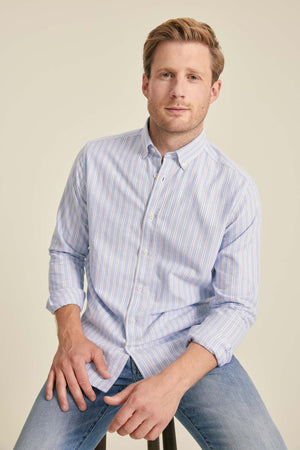 Man wearing a light blue striped shirt and jeans sitting on a stool against a beige background