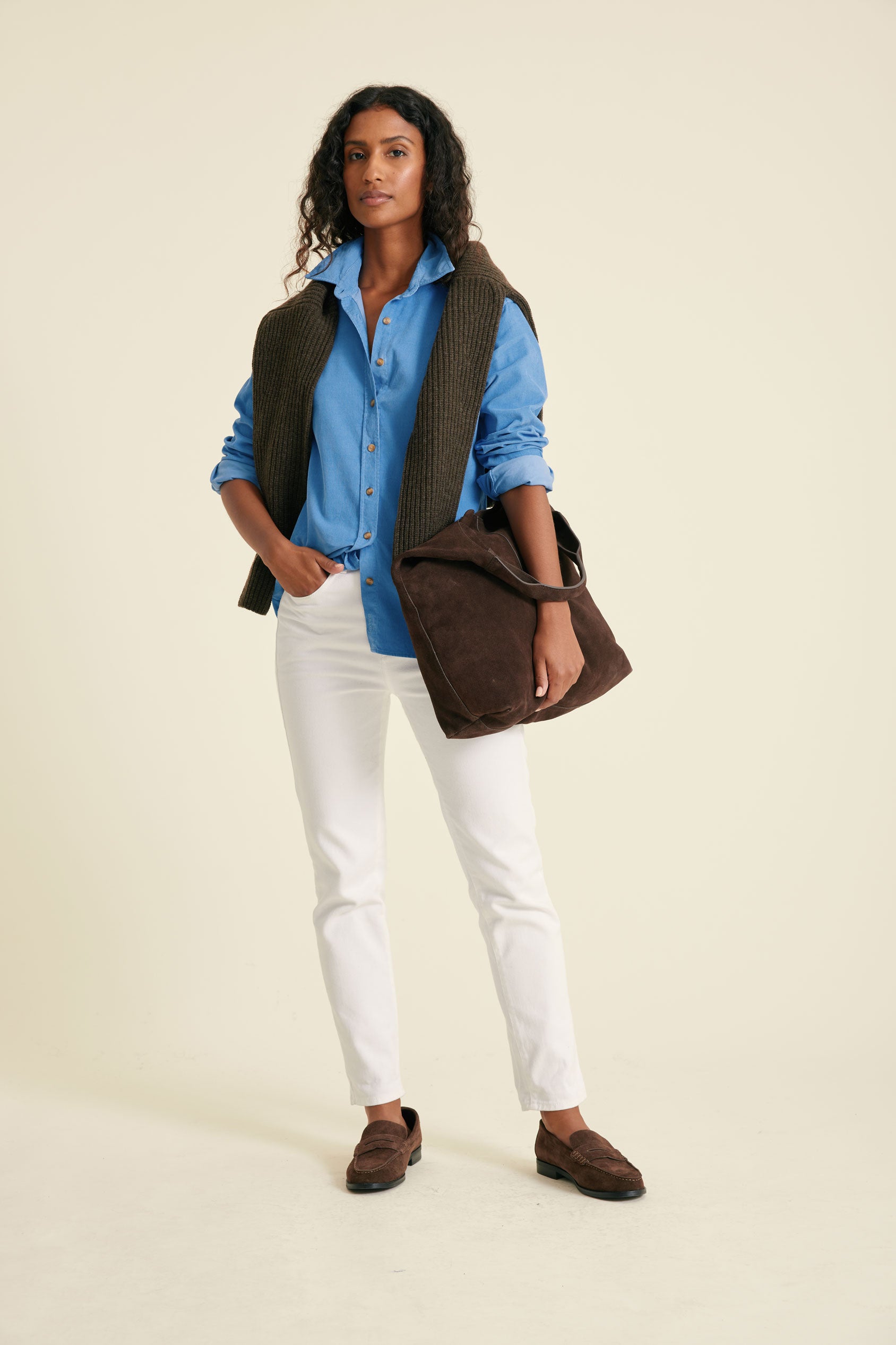 Woman wearing a blue shirt, white pants, brown vest, and carrying a brown bag on a beige background