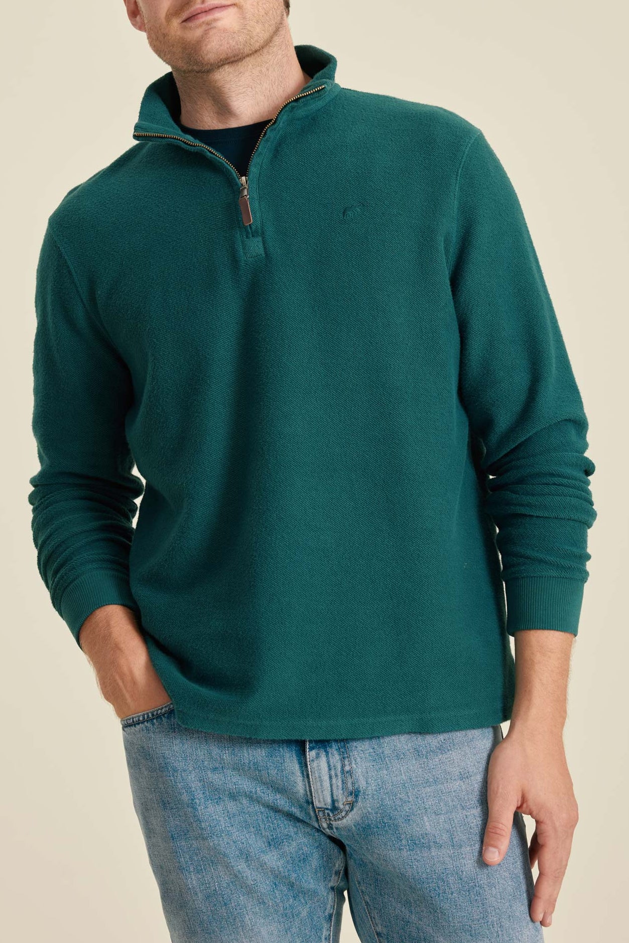 Man wearing a teal fleece pullover and jeans on a beige background